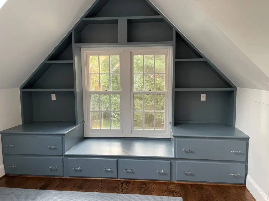 Built-in window seat with drawers and shelving under a gable window, painted blue-gray