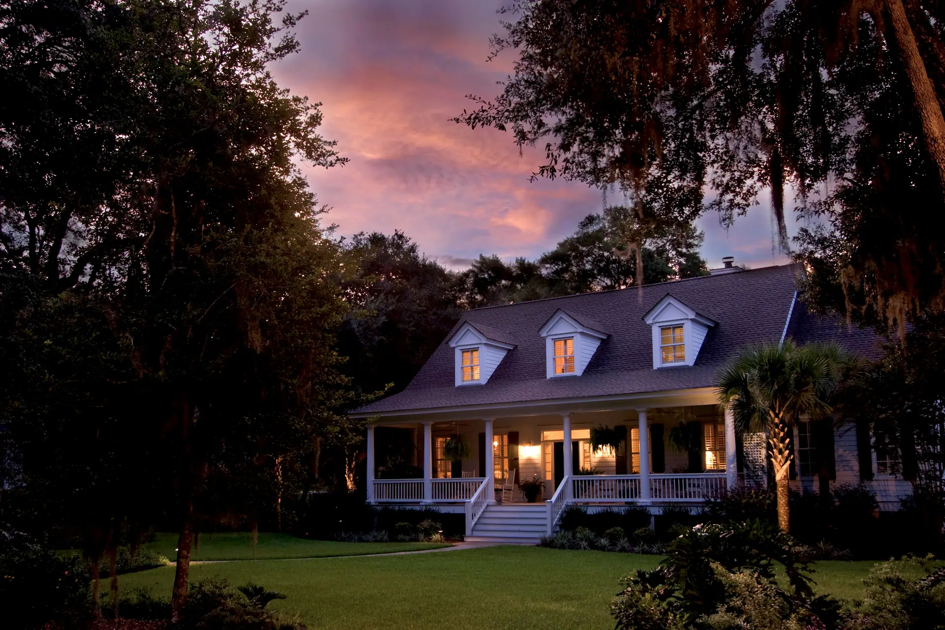 Two-story home with dormers and a lit wraparound front porch at dusk