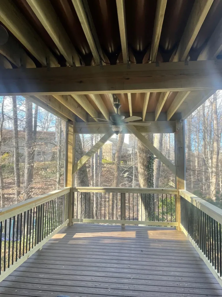Covered porch under an upper deck, with a ceiling fan and black railings looking out to trees